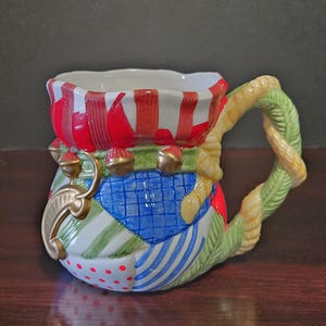 May include: A decorative ceramic mug with a patchwork design. The mug features red and white stripes, green and blue patterns, and gold accents. The handle is a twisted rope design with green leaves.