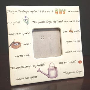 May include: White ceramic picture frame with a quote about gentle drops replenishing the earth and renewing our spirit. The frame has a small opening for a photo and features illustrations of plants, a bird's nest, a bee, and a watering can.