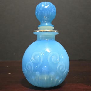 Puede incluir: Una botella de vidrio azul claro con un diseño de espiral y un tapón azul a juego. La botella tiene una banda plateada alrededor del cuello.