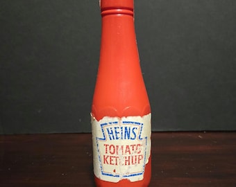 May include: A bright orange plastic toy bottle with a screw-top cap. A white paper label is wrapped around the lower section, featuring a blue border and the printed text HEINS TOMATO KETCHUP in blue and red lettering with a worn, aged appearance.