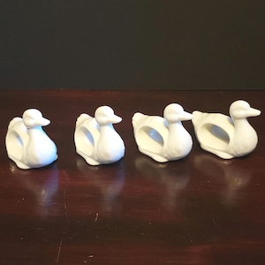 May include: Set of four white ceramic duck napkin rings. The ducks are in a row, facing the right side of the image.