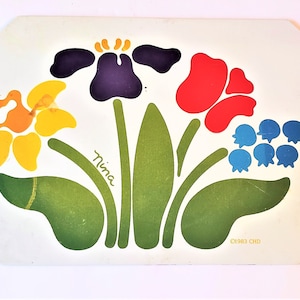 May include: A white ceramic trivet with a colorful floral design. The design features a purple iris, a red poppy, a yellow daffodil, and bluebells. The trivet is signed "Nina" and dated "1983 CHD".