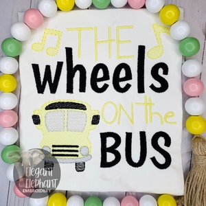 May include: A white t-shirt with yellow and black embroidery. The embroidery depicts a school bus with the text "THE wheels ON the BUS" and musical notes.