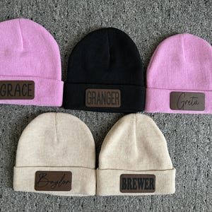 May include: Five knit beanies with leather patches. The beanies are in pink, black, and beige. The patches have the names "Grace", "Granger", "Greta", "Baylor", and "Brewer" written on them.