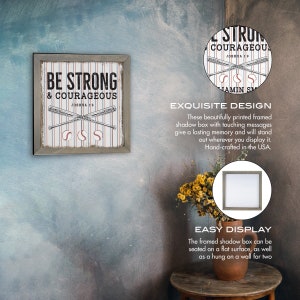 May include: A framed wood shadow box with a white and red striped background, two crossed baseball bats, and three baseballs. The text "BE STRONG & COURAGEOUS" is printed in black with the scripture reference "JOSHUA 1:9" below. The shadow box is hanging on a light blue wall.