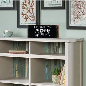 May include: A black wooden sign with white text that reads "I just want to sip coffee & pet my cat". The sign has a small coffee cup icon and a paw print. The sign is sitting on a white shelf with other items.