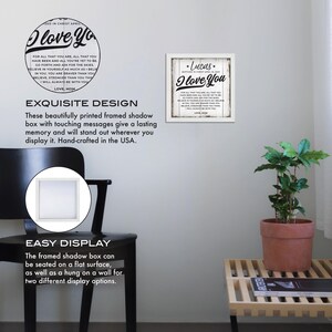 May include: A white framed shadow box with a black and white print that reads "I love you" with a personalized name and date. The print is surrounded by a black border and has a distressed white background. The shadow box is displayed on a white wall with a black chair and a potted plant in the background.