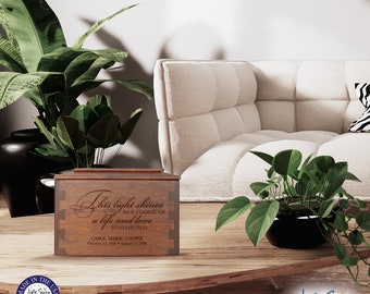 May include: A wooden box with a personalized inscription that reads "This light shines as a symbol of a life and love remembered. Carol Marie Cooper, October 22, 1958 - January 2, 2018". The box is sitting on a wooden table with a green plant in a black pot.