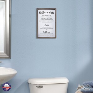 May include: A framed sign with the text "Bathroom Rules" and a list of rules for using the bathroom. The sign is hanging on a light blue wall in a bathroom.