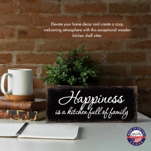 May include: Black wooden sign with white text that reads "Happiness is a kitchen full of family". The sign is sitting on a white surface with a book and a mug.