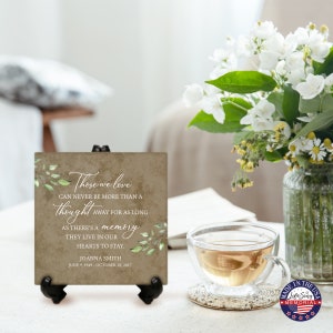 May include: A brown and green ceramic tile with a quote about love and memory. The quote reads "Those we love can never be more than a thought away for as long as there's a memory they live in our hearts to stay." The tile also includes the name Joanna Smith and the dates June 9, 1949 - October 18, 2017. The tile is on a black stand.