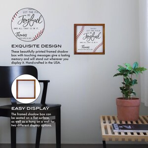 May include: A framed shadow box with a baseball-themed design. The box features a red and white baseball with stitching, and the text "Let the field be joyful and all that is in it. Thomas." The box is displayed on a white wall with a plant in a terracotta pot on a wooden table.