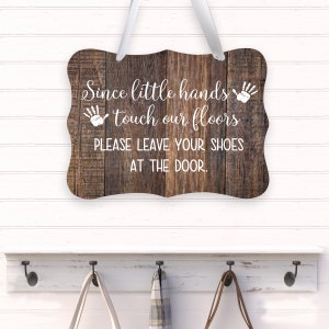May include: Wooden sign with the text "Since little hands touch our floors PLEASE LEAVE YOUR SHOES AT THE DOOR." The sign is brown with white text and a white ribbon for hanging. A white shelf with hooks is below.