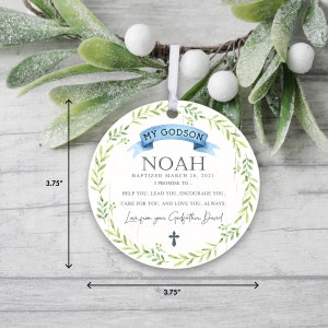 May include: A round, white ornament with a green leafy border. The ornament has a blue ribbon with the text "My Godson Noah Baptized March 20, 2021 I Promise To... Help you, lead you, encourage you, care for you, and love you, always. Love from your Godfather, David" and a small cross.