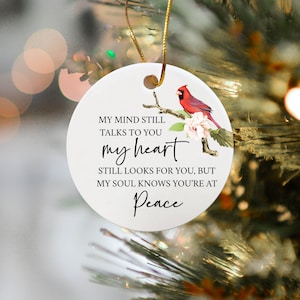 May include: A white round ornament with a red cardinal bird perched on a branch with white flowers. The ornament has the text "MY MIND STILL TALKS TO YOU my heart STILL LOOKS FOR YOU, BUT MY SOUL KNOWS YOU'RE AT Peace".