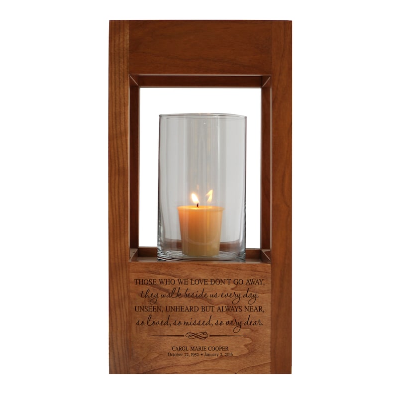 May include: A wooden candle lantern with a glass cylinder and a lit candle inside. The lantern has an engraved message that reads: "Those who we love don't go away, they walk beside us every day. Unseen, unheard but always near, so loved, so missed, so very dear." The lantern also has the name "CAROL MARIE COOPER" and the dates "October 22, 1952 - January 2, 2006" engraved on it.