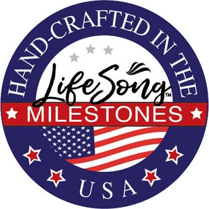 Pu&ograve; includere: Un cerchio blu con testo bianco che recita "Hand-Crafted in the USA". All'interno del cerchio c'&egrave; una bandiera americana rossa, bianca e blu con il testo "Life Song Milestones" in nero.