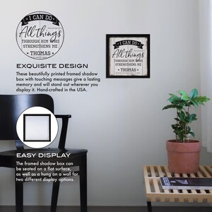 May include: A framed shadow box with a white background and black text. The text reads "I can do all things through him who strengthens me - Thomas". The box is displayed on a white wall with a plant in a terracotta pot on a wooden shelf.