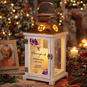 Puede incluir: Linterna de madera blanca con tapa y asa de cobre. La linterna presenta un diseño de colibrí y flores con el texto "Hummingbirds appear with angels are near". Una foto enmarcada y piñas se encuentran en el fondo.
