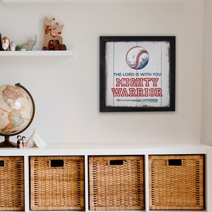 May include: Framed baseball-themed wall art with a black frame. The artwork features a baseball with a mountain scene and the text "THE LORD IS WITH YOU MIGHTY WARRIOR" in red and blue. A baseball bat graphic is below the text.