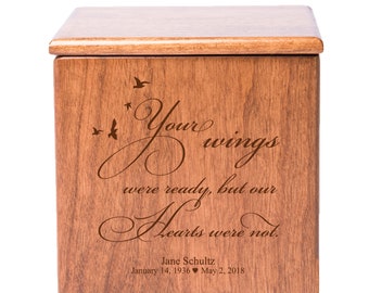 May include: A wooden cremation urn with a rectangular shape and a light brown finish. The front of the urn features engraved text that reads "Your wings were ready, but our hearts were not." Below the text are the name "Jane Schultz" and dates.