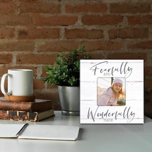 May include: A white wooden photo frame with the words "Fearfully and Wonderfully Made" in gray script. The frame holds a photo of a young girl. A white mug, books, and a plant are also on the table.