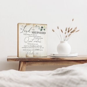 May include: A wooden sign with a rustic background and green foliage accents. The sign features a bible verse from Psalm 23, "The Lord is my Shepherd, I lack nothing. He makes me lie down in green pastures, He leads me beside quiet waters. He refreshes my soul... Surely Your Goodness and Love will follow me all the days of my life, and I will dwell in the house of the Lord forever. Psalm 23"