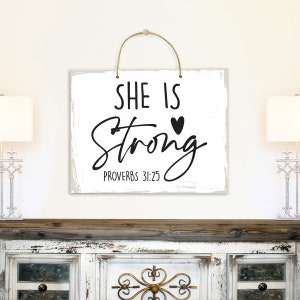 May include: White wooden sign with black text that reads "She is Strong" with a small heart symbol and "Proverbs 31:25" below.