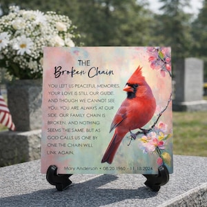 May include: A memorial plaque with the text "The Broken Chain" and a red cardinal bird illustration. The plaque includes the name Mary Anderson and dates 08.20.1960 - 11.18.2024. The background has a soft, watercolor-like effect with floral accents.