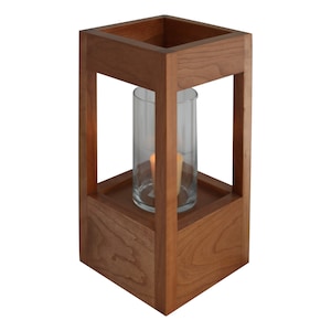 May include: A wooden candle lantern with a clear glass cylinder and a lit candle inside. The lantern is made of cherry wood and has a square base.