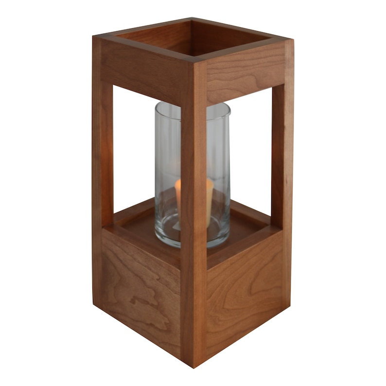 May include: A wooden candle lantern with a clear glass cylinder and a lit candle inside. The lantern is made of cherry wood and has a square base.