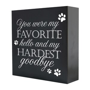 May include: Black wooden sign with white text that reads "You were my FAVORITE hello and my HARDEST goodbye" with paw prints on either side of the text.