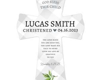 May include: A white cross-shaped keepsake with a gray gingham pattern. The cross features the text "GOD BLESS THIS CHILD," the name "LUCAS SMITH," and the date "04.16.2023." Additional text includes a Bible verse and a sprig of leaves.
