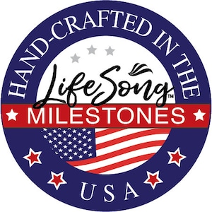 May include: A blue circle with white text that reads "Hand-Crafted in the USA". Inside the circle is a red, white, and blue American flag with the text "Life Song Milestones" in black.