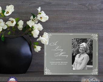 May include: A gray picture frame with a white border and a white inscription that reads "In Loving Memory". The frame has a photo of a woman smiling. The inscription below the photo reads "Lydia Kate Wilson May 6, 1957 - August 15, 2009".