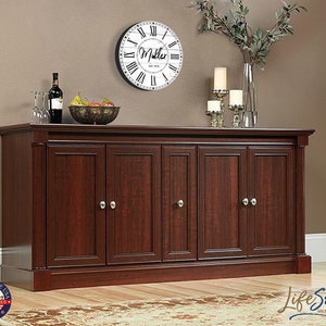 May include: A dark wood cabinet with six doors and silver door knobs. The cabinet is sitting on a wooden floor with a rug in front of it. There is a clock on the wall behind the cabinet with the name "Miller" on it.