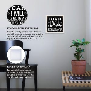 May include: A black and white framed shadow box with the text "I can I will I believe whatever it takes I'll never give up". The box is displayed on a white wall with a black chair and a potted plant in the background.