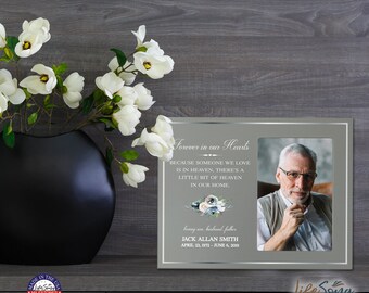 May include: A memorial photo frame with a picture of an elderly man, text that reads "Forever in our Hearts", and floral accents. A black vase with white flowers is to the left. The frame includes the name Jack Allan Smith and dates.
