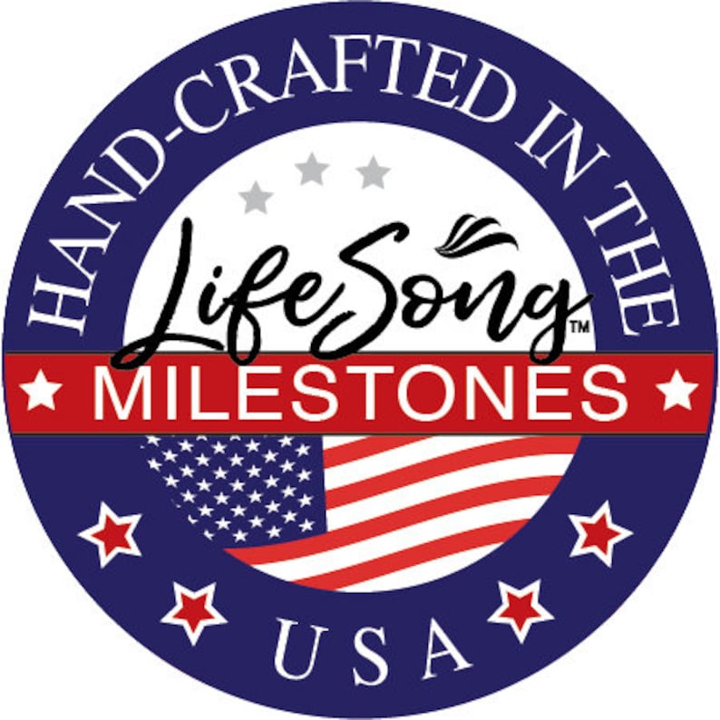 May include: A blue circle with white text that reads "Hand-Crafted in the USA". Inside the circle is a red, white, and blue American flag with the text "Life Song Milestones" in black.