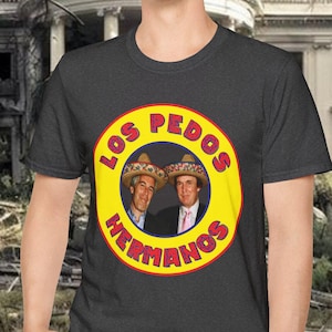 May include: A dark gray t-shirt featuring a yellow circle with the words "LOS PEDOS HERMANOS" in red. Inside the circle are two men wearing sombreros. The background shows a damaged building.