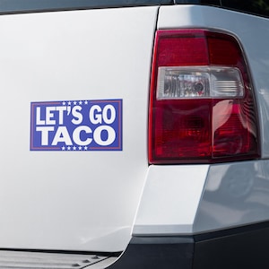 May include: A white vehicle with a rectangular bumper sticker that reads "LET'S GO TACO" in white text on a blue background. The sticker is on the rear of the vehicle, next to a red tail light.
