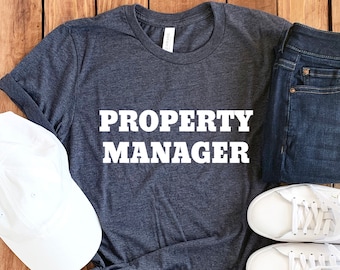 May include: A dark gray t-shirt with the text "PROPERTY MANAGER" in white letters. The shirt is laid out on a wooden surface with a white baseball cap, a pair of blue jeans, and two white sneakers.
