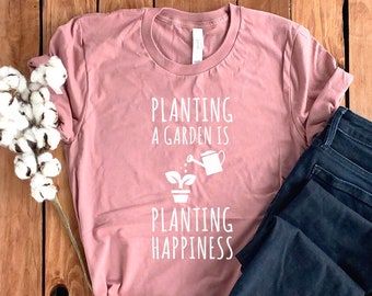 May include: A dusky pink t-shirt with the words "Planting a garden is planting happiness" in white. The shirt is next to dark blue jeans and a sprig of cotton on a wooden surface.