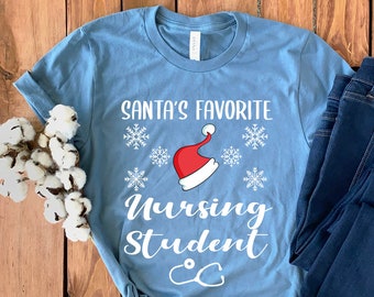 Könnte beinhalten: Ein blaues T-Shirt mit weißem Text, der "Santa's Favorite Nursing Student" mit einem roten Weihnachtsmann-Hut und Schneeflocken sagt. Das Shirt liegt auf einer Holzoberfläche mit einer blauen Jeans.