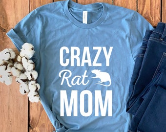May include: A light blue t-shirt with the text "CRAZY Rat MOM" in white lettering. A white rat silhouette is next to the word "Rat". The t-shirt is laid out on a wooden surface with a pair of blue jeans.