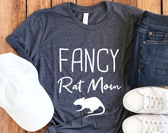 May include: A dark gray t-shirt with the text "FANCY Rat Mom" in white lettering and a white silhouette of a rat. The shirt is laid out on a wooden surface with a white baseball cap, a pair of blue jeans, and a pair of white sneakers.