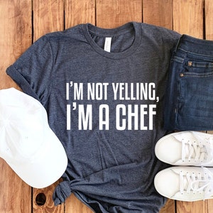 May include: A dark grey t-shirt with the text "I'm not yelling, I'm a chef" printed in white. The shirt is laid out on a wooden surface with a white baseball cap, a pair of blue jeans, and two white sneakers.
