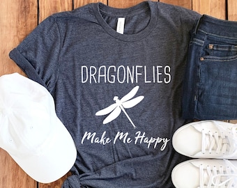 May include: A dark gray t-shirt with the words "DRAGONFLIES Make Me Happy" and a dragonfly graphic. The shirt is paired with blue jeans, white sneakers, and a white baseball cap, all arranged on a wooden surface.