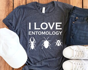 May include: A dark gray t-shirt with the text "I Love Entomology" and images of insects. The shirt is laid out on a wooden surface with a white baseball cap, a pair of blue jeans, and two white sneakers.