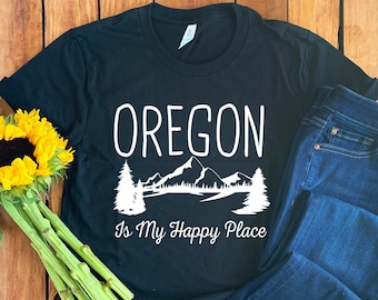 May include: Black t-shirt with white text that reads "OREGON Is My Happy Place" and a graphic of mountains and trees. The shirt is laid out on a wooden surface with a pair of blue jeans and a bouquet of sunflowers.